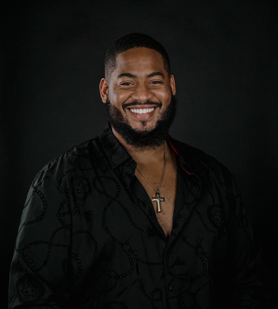 Tiye Martin smiling, in a dark patterned shirt and a cross necklace, against a black background